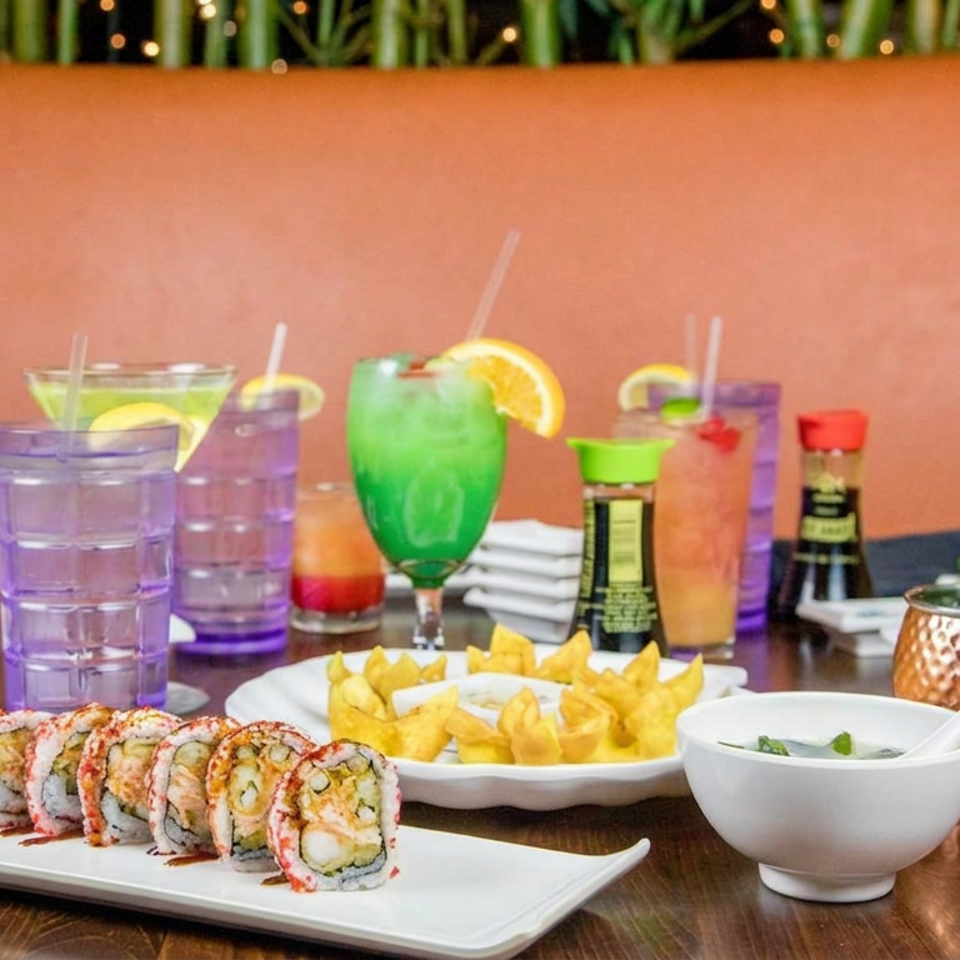 Sushi rolls, golden fries, colorful cocktails, dipping sauces, and condiments fill a restaurant table against a warm orange banquette.