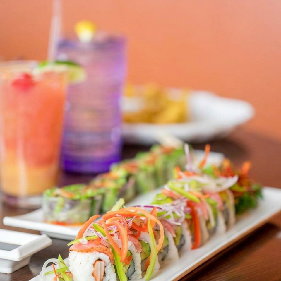 Colorful sushi rolls topped with shredded vegetables alongside dipping sauce and tropical drinks on a restaurant table.