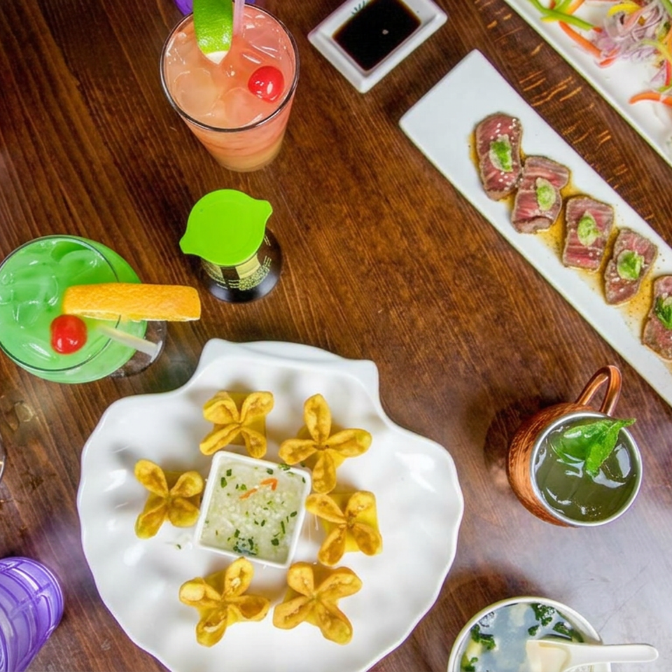Overhead view of colorful cocktails, flower-shaped appetizers with dipping sauce, sliced steak rolls, and vegetable salad on a wooden table.