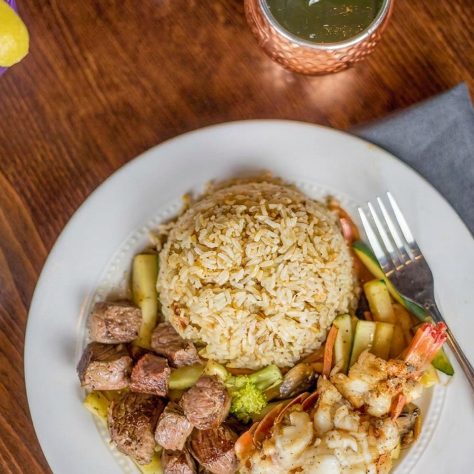 Plate of seasoned rice, grilled steak bites, shrimp, and vegetables on a wooden restaurant table with drinks.