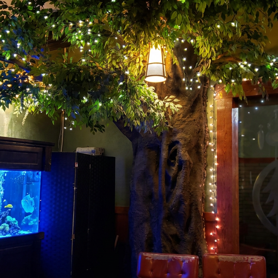 Cozy restaurant interior featuring a brightly lit aquarium, artificial tree with string lights, pendant lighting, and wooden entrance doors.