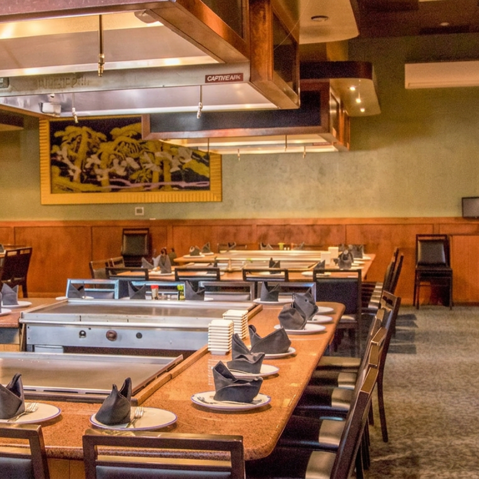 Spacious Japanese teppanyaki restaurant dining room with grill tables, folded napkins, wooden furnishings, and overhead ventilation hoods.